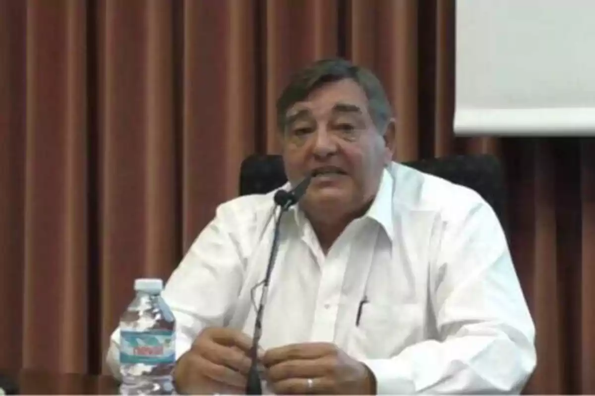 Hombre mayor con camisa blanca hablando en un micrófono durante una conferencia, con una botella de agua sobre la mesa y cortinas marrones de fondo.
