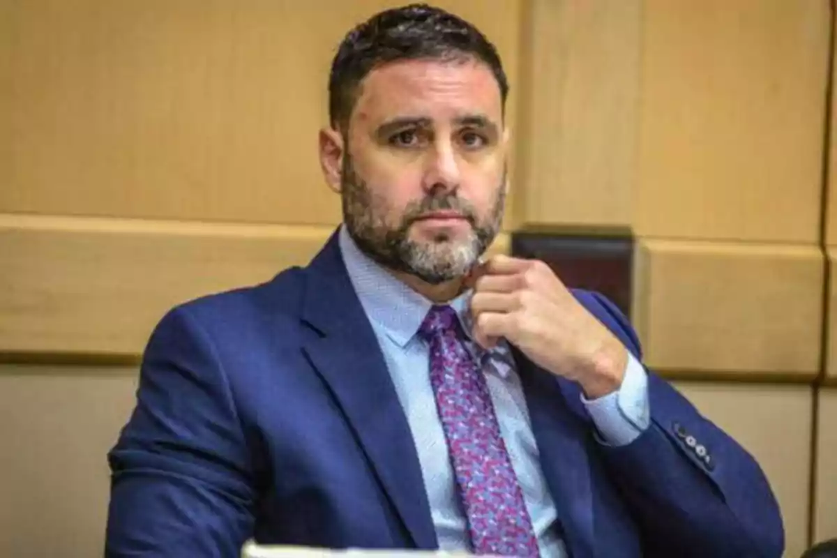 Hombre con traje azul y corbata morada sentado en una sala de conferencias.