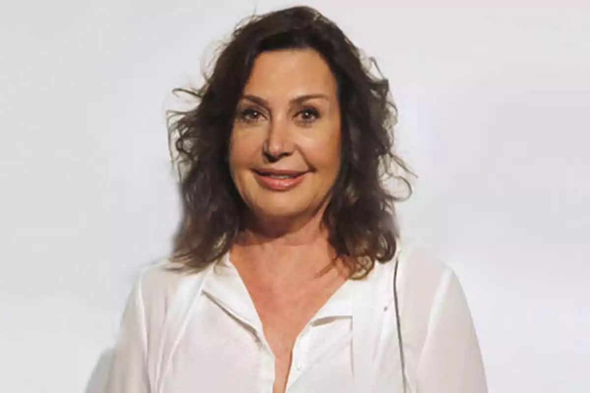 Mujer con cabello oscuro y rizado, sonriendo, vestida con una blusa blanca, sobre un fondo claro.