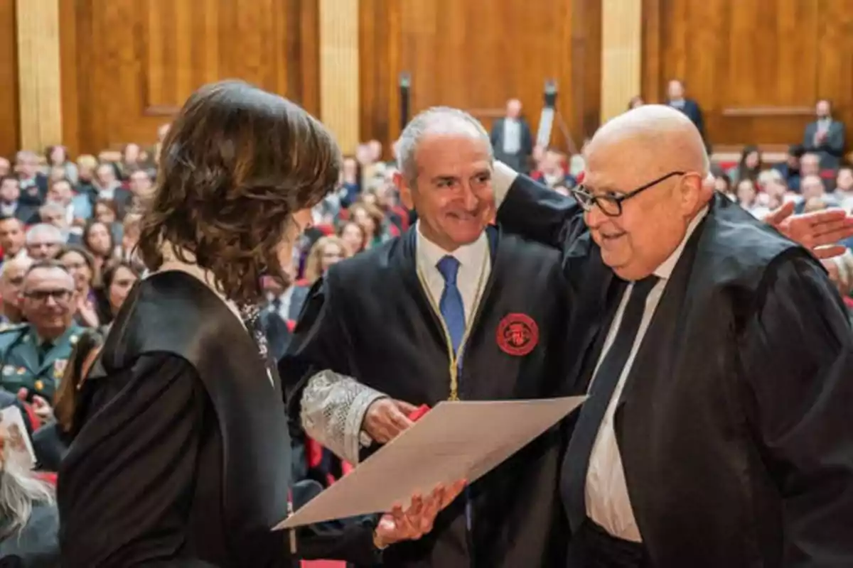 Tres personas con togas académicas están en una ceremonia, una de ellas sostiene un documento mientras las otras dos sonríen y se miran, en el fondo se puede ver una audiencia sentada en un salón con paredes de madera.