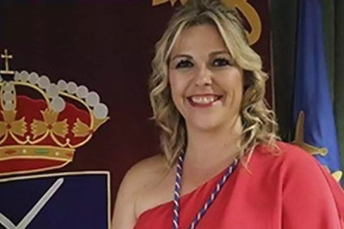 Mujer rubia sonriendo con un vestido rojo frente a un fondo con un escudo.