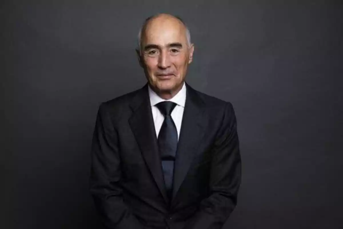 Hombre mayor con traje oscuro y corbata, posando frente a un fondo gris.