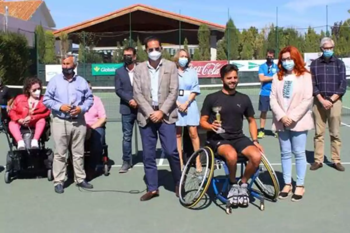 Un grupo de personas, algunas en sillas de ruedas, posan en una cancha de tenis al aire libre, con una persona sosteniendo un trofeo en el centro.