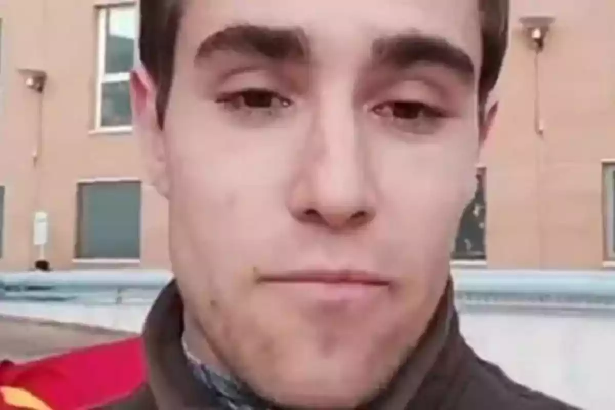 Un hombre joven con cabello corto y oscuro, vestido con una chaqueta, se encuentra al aire libre frente a un edificio de ladrillo con ventanas.