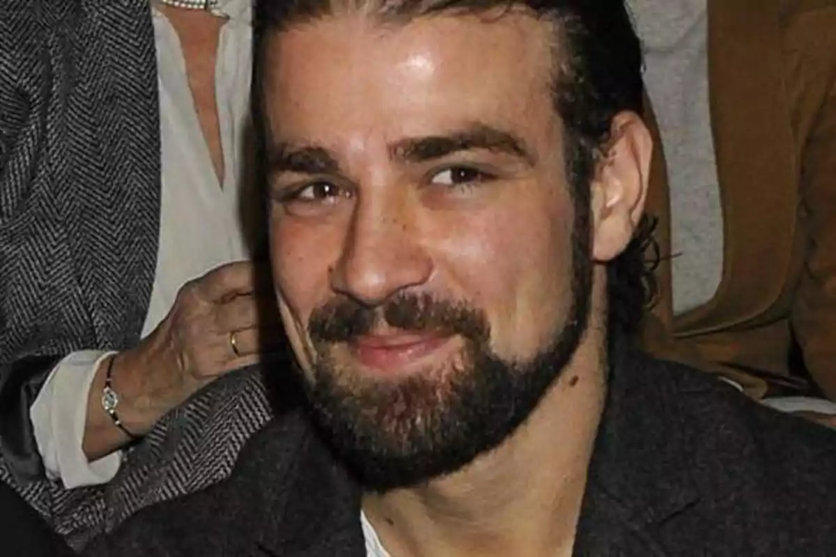 Un hombre con barba y cabello oscuro sonríe mientras está sentado, con personas en el fondo.