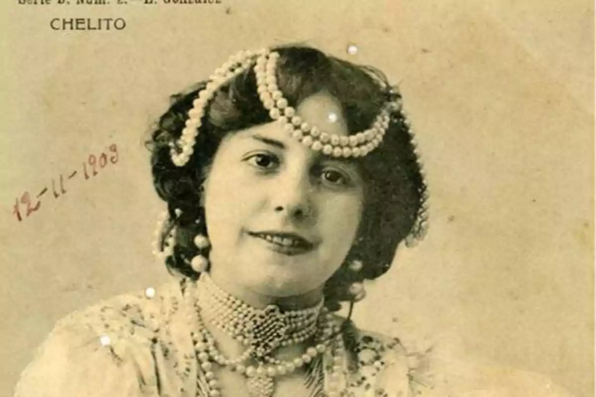 Una mujer con un peinado adornado con perlas y un collar de perlas, vestida con ropa elegante, en una fotografía antigua fechada el 12-11-1908.