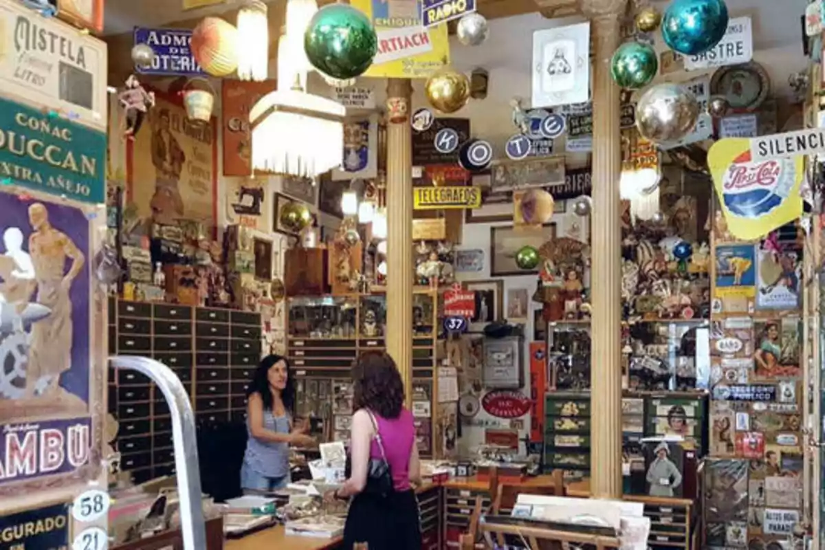 Una tienda de antigüedades llena de carteles vintage, objetos decorativos y dos personas conversando en el mostrador.