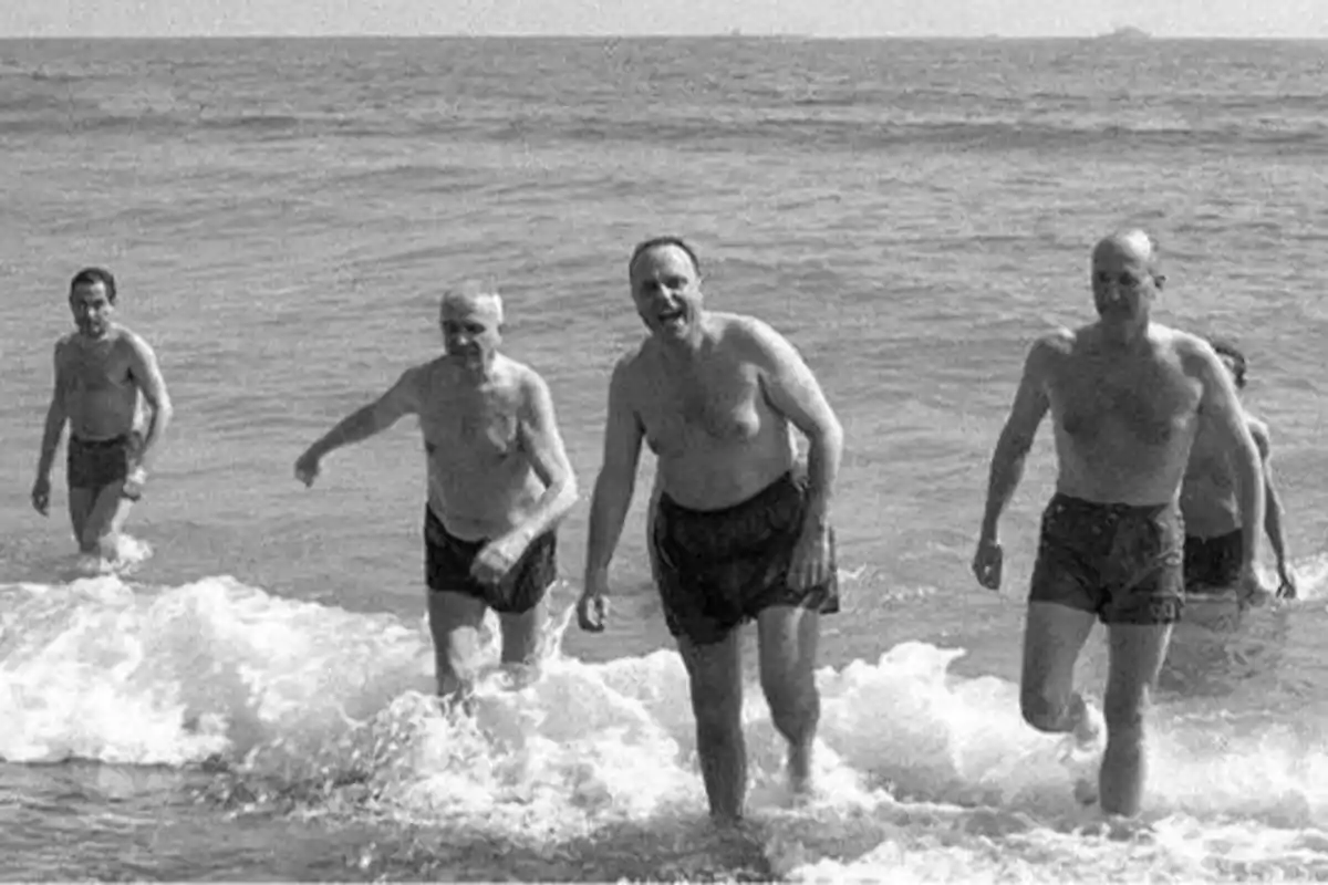 Cinco hombres en traje de baño salen del mar caminando hacia la orilla.