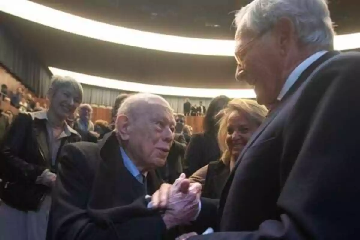 Dos hombres mayores se saludan con un apretón de manos en un evento, mientras otras personas observan y sonríen en el fondo.