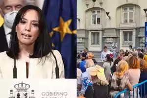 Una mujer hablando en una conferencia de prensa junto a una imagen de una multitud reunida frente a un edificio.