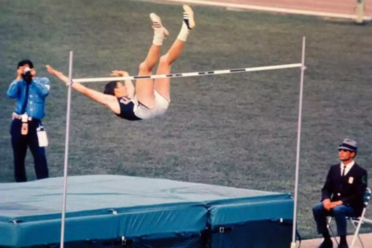 Atleta realizando un salto de altura en una competencia, mientras un fotógrafo y un juez observan.