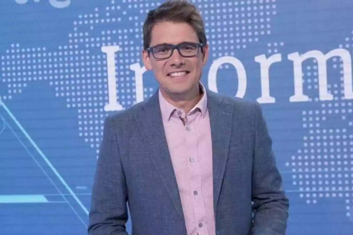 Un hombre con gafas y una chaqueta gris sonríe frente a un fondo azul con texto parcialmente visible.