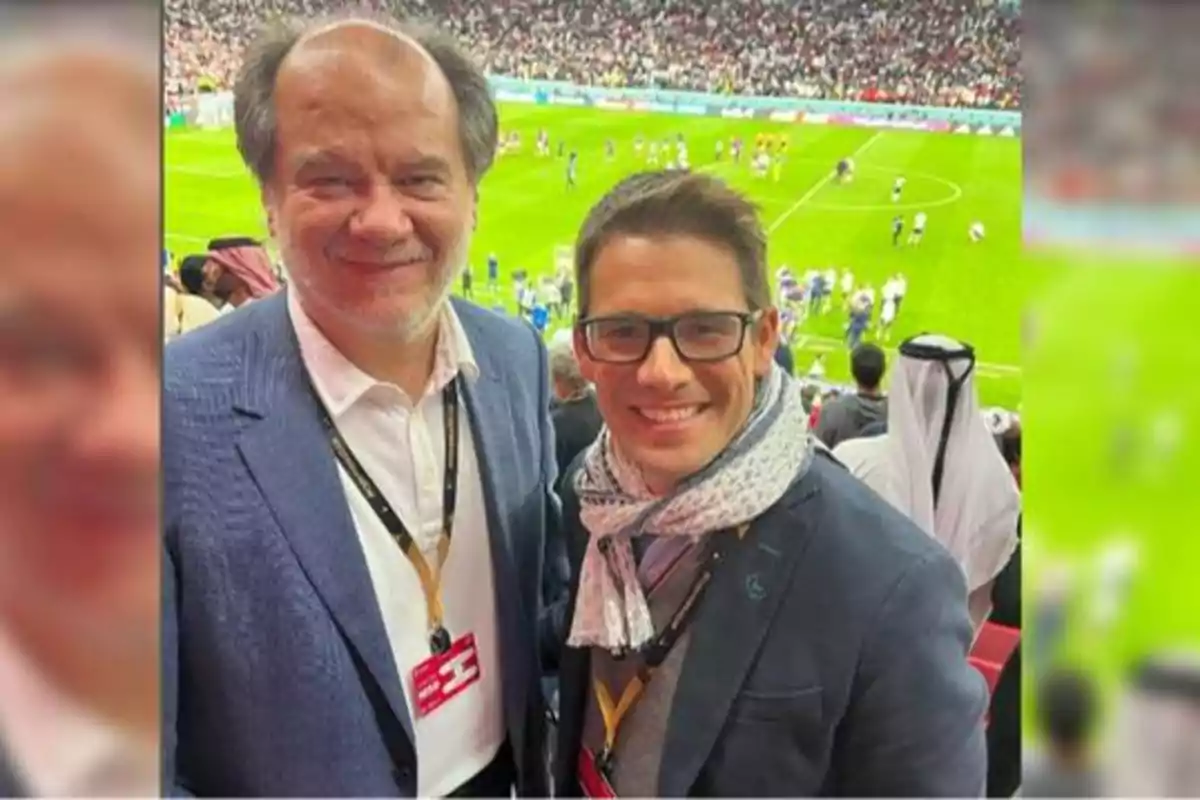 Dos personas sonrientes posan para una foto en un estadio de fútbol lleno de espectadores, ambos llevan gafetes colgados al cuello y visten ropa formal.