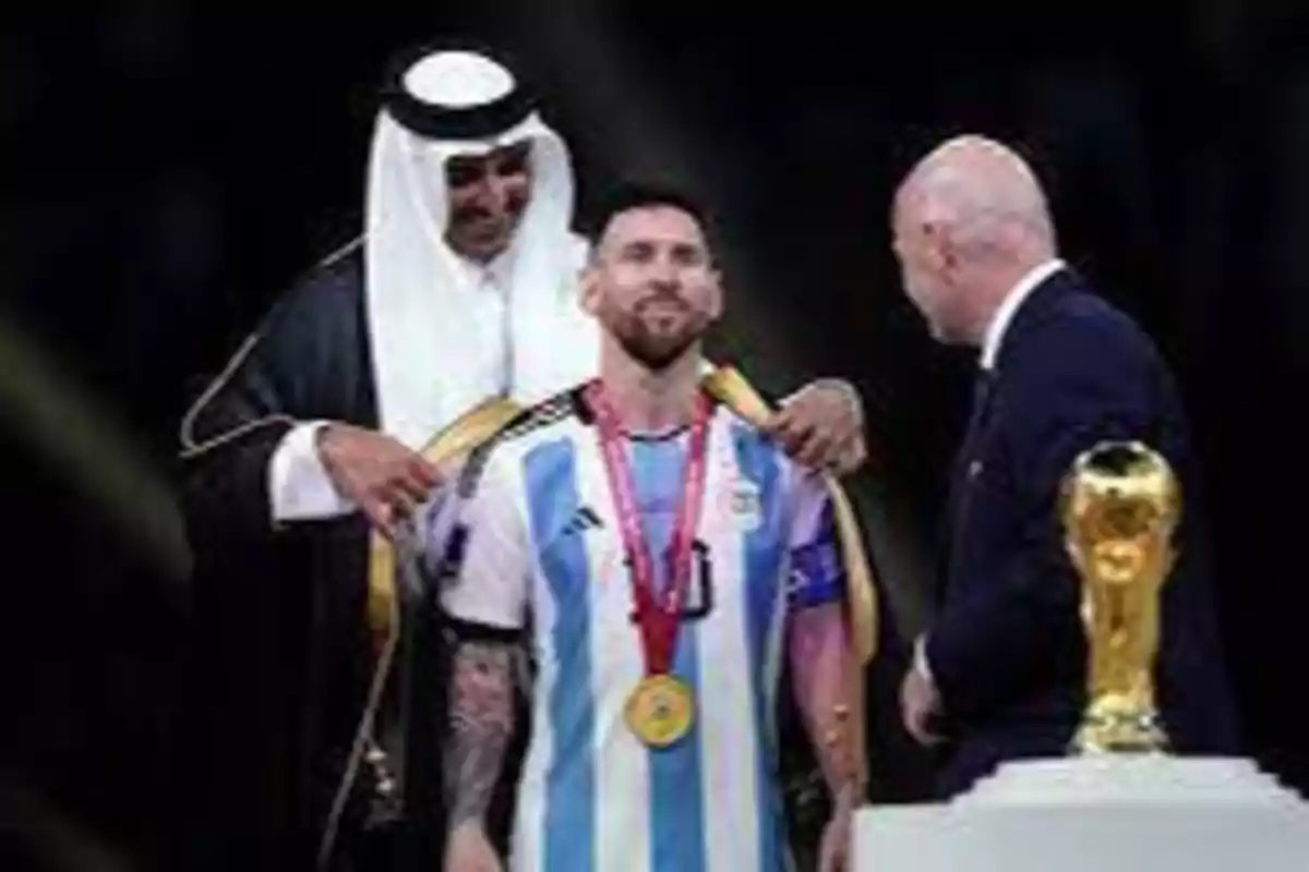 Un hombre con una túnica blanca coloca una capa sobre los hombros de un futbolista con la camiseta de Argentina, mientras otro hombre calvo con traje oscuro observa; en primer plano se ve un trofeo dorado.