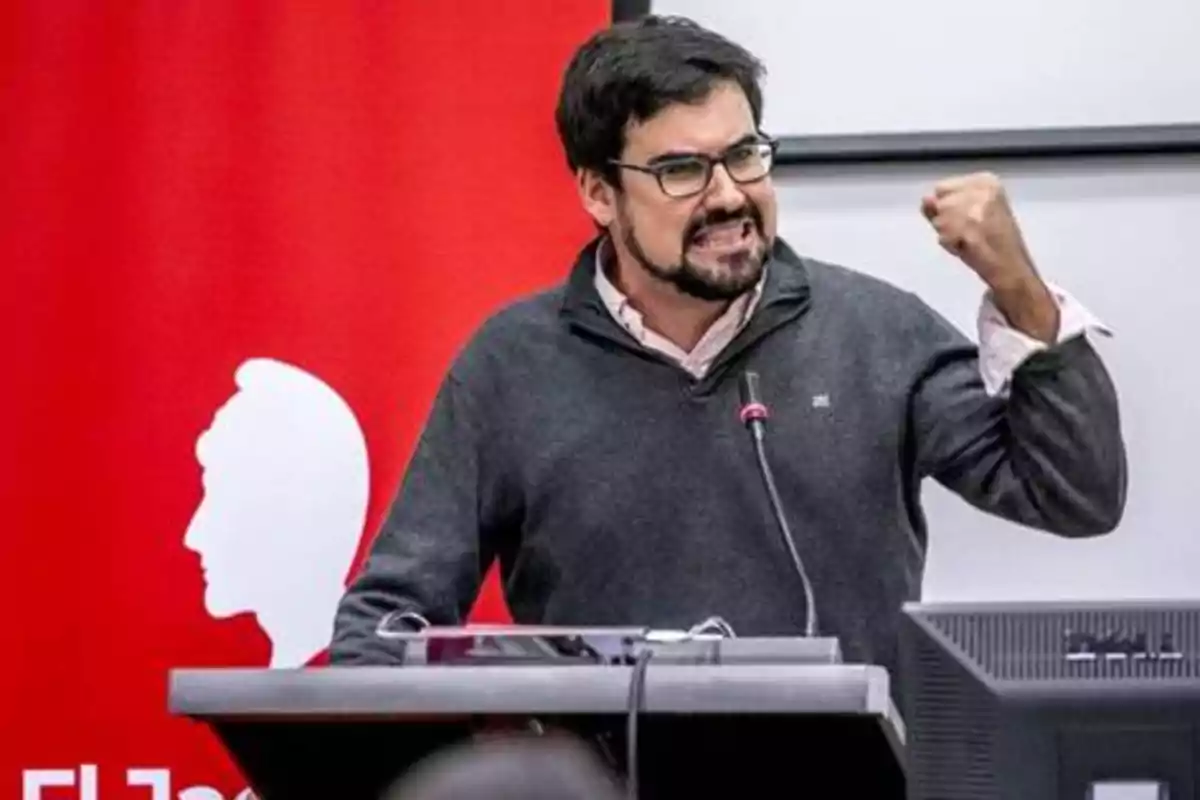 Hombre con gafas y barba gesticulando con el puño levantado mientras habla en un podio con un micrófono, fondo rojo con silueta de perfil.