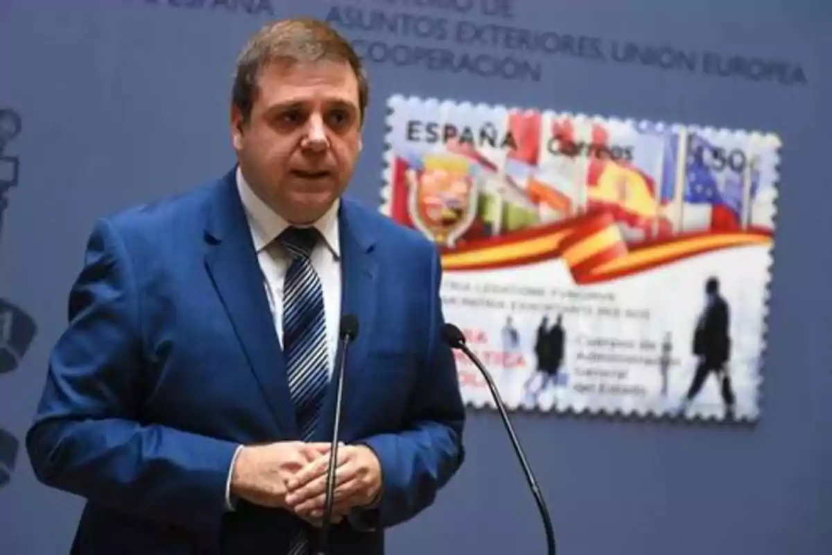 Hombre en traje azul hablando en un podio con un fondo que muestra un sello postal de España.