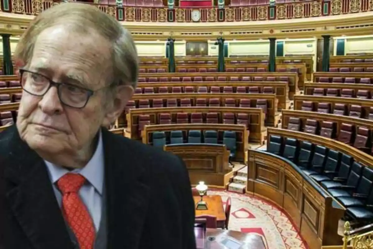 Un hombre mayor con gafas y corbata roja está en primer plano, con un parlamento vacío de fondo.