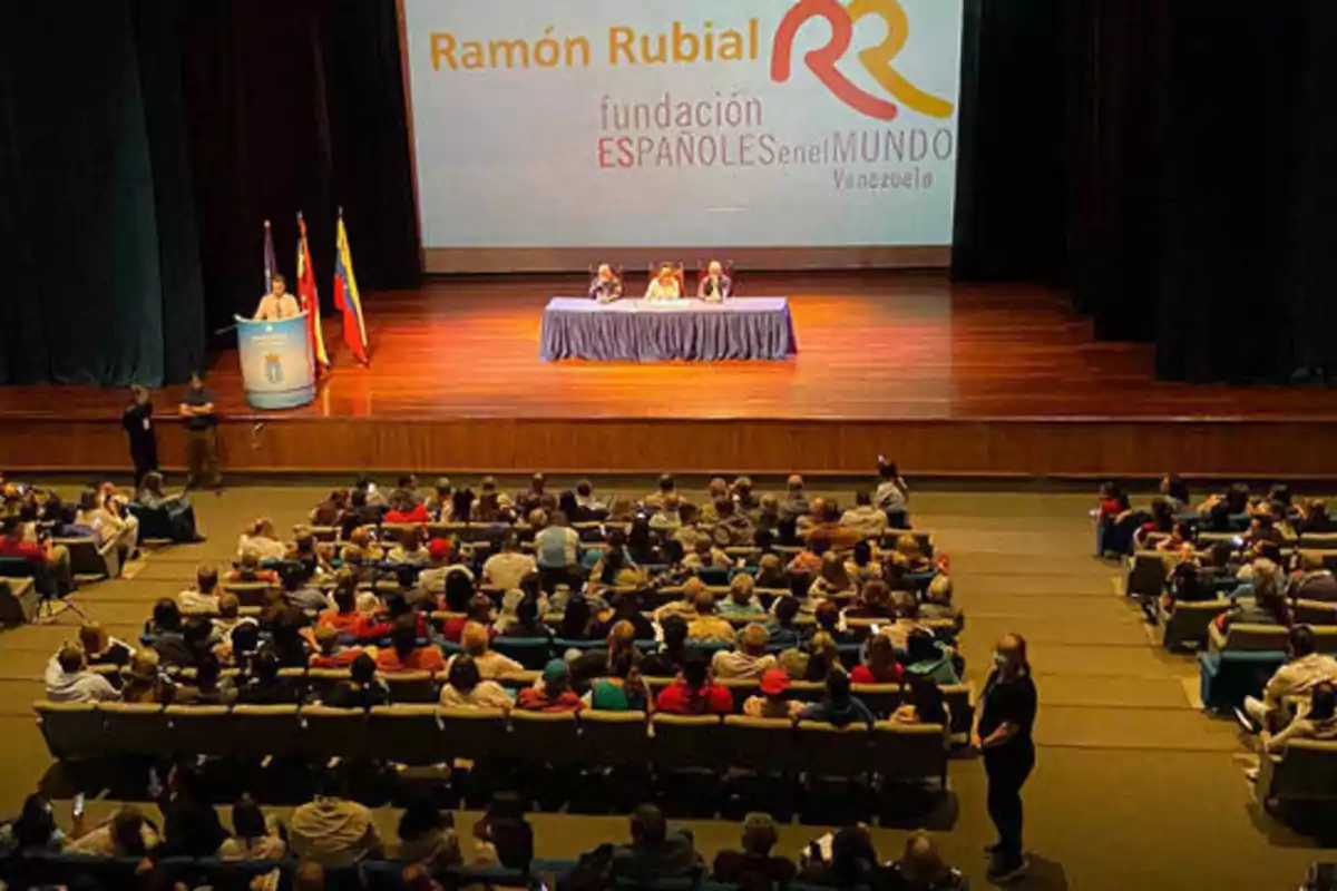 Una conferencia de la Fundación Españoles en el Mundo en Venezuela, con un auditorio lleno de asistentes y un escenario con ponentes y banderas.