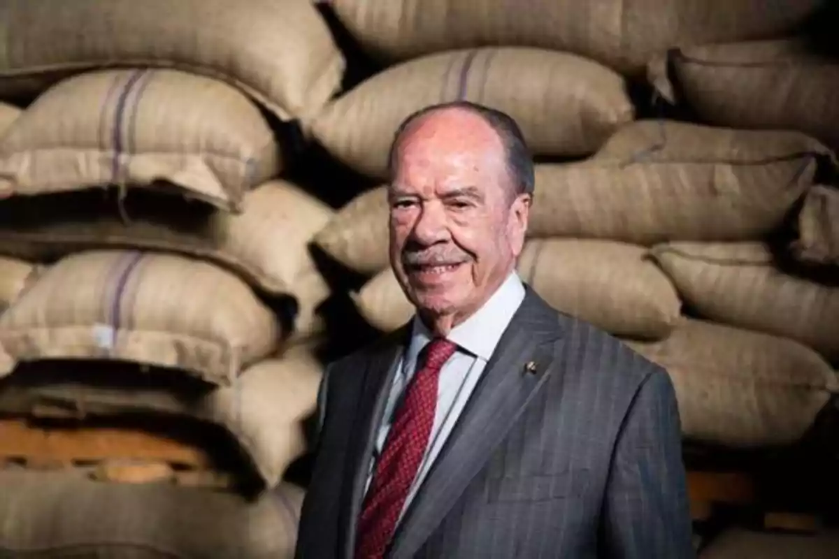 Hombre mayor con traje y corbata roja sonriendo frente a una pila de sacos de yute.