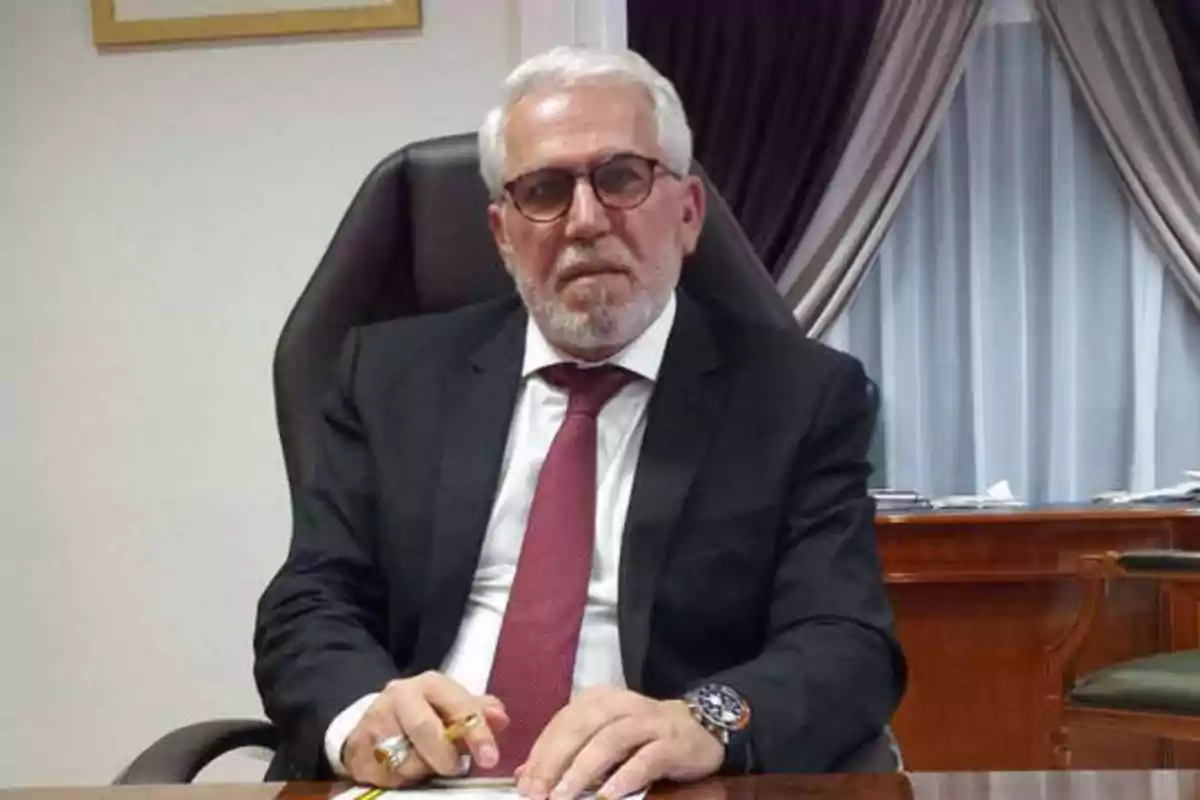 Un hombre de cabello canoso y barba, con gafas y traje formal, sentado en un escritorio con cortinas de fondo.