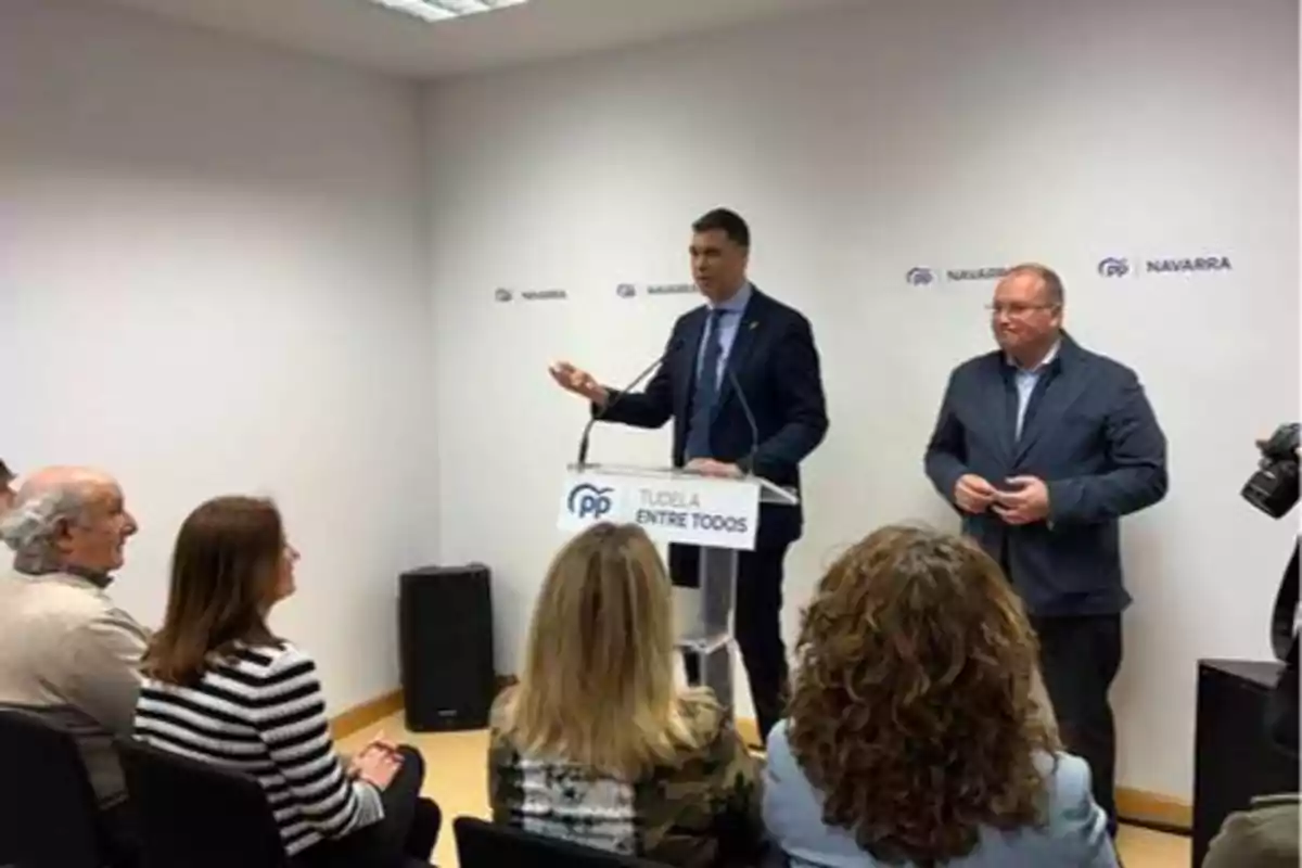 Un hombre de traje habla en un podio con el logo del PP y el texto "TUDELA ENTRE TODOS" mientras otro hombre está de pie a su lado y varias personas están sentadas frente a ellos en una sala con paredes blancas y el logo de Navarra en el fondo.