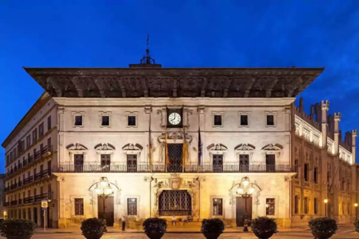 Edificio histórico iluminado por la noche con un reloj en el centro y un cielo azul de fondo.