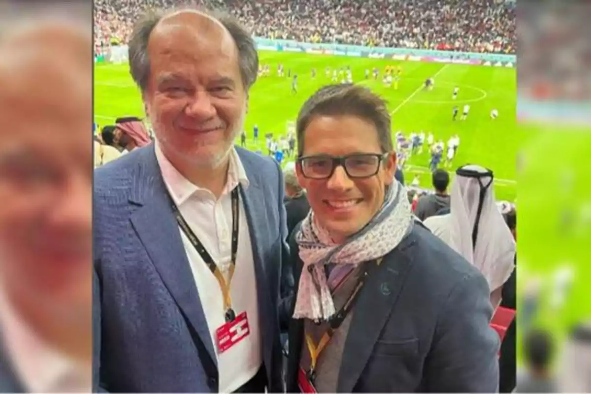 Dos personas posan para una foto en un estadio de fútbol lleno de espectadores, ambos llevan gafetes colgados al cuello y visten ropa formal.