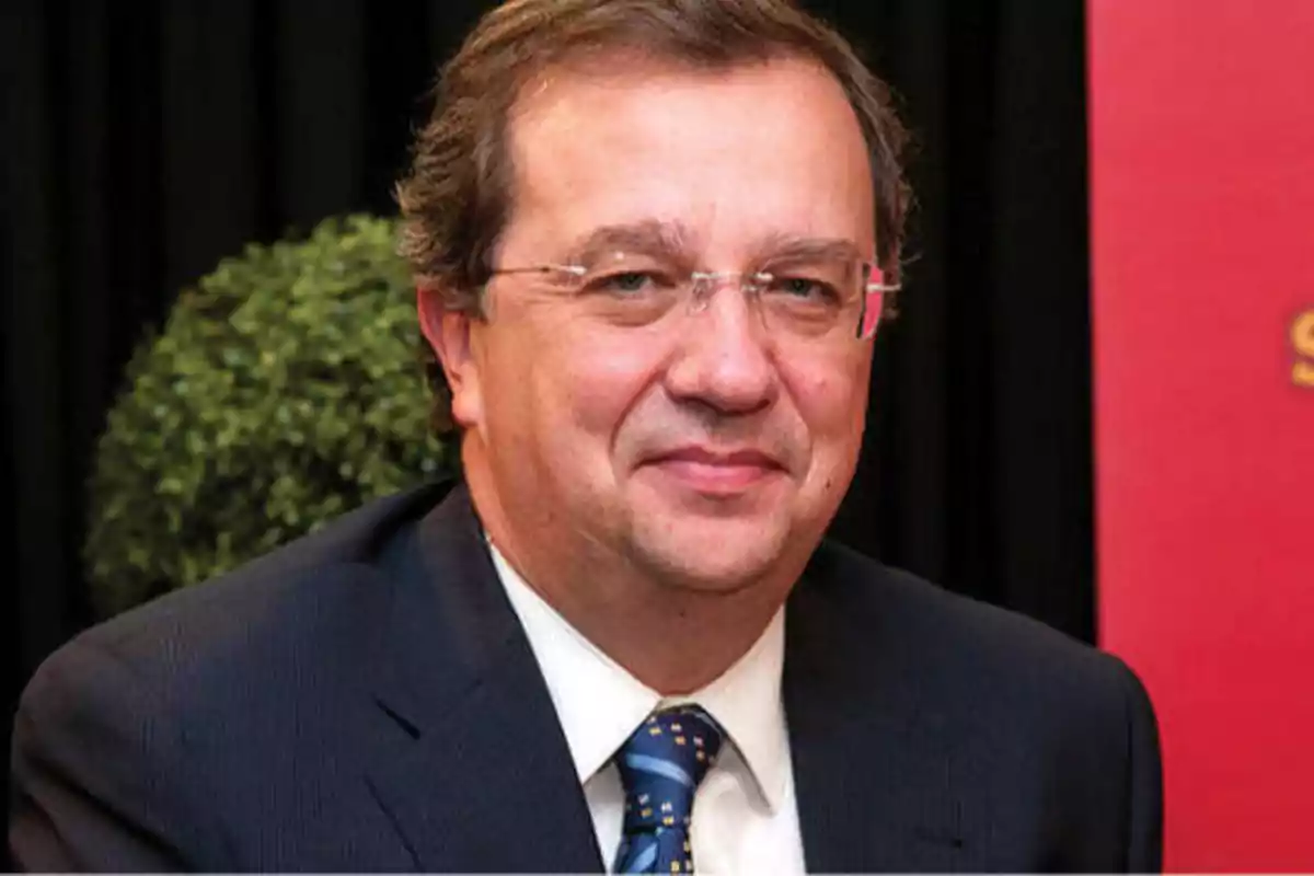 Hombre con gafas y traje oscuro sonriendo frente a un fondo negro y rojo.