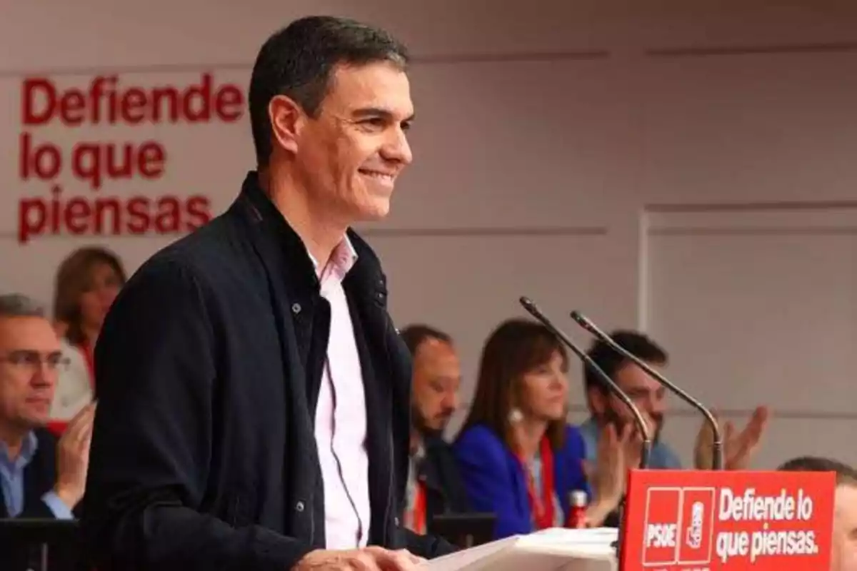 Un hombre hablando en un podio con un cartel que dice "Defiende lo que piensas" y el logotipo del PSOE, con varias personas sentadas al fondo.