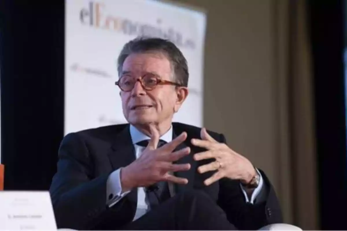 Un hombre con gafas y traje oscuro gesticulando mientras habla en un evento.