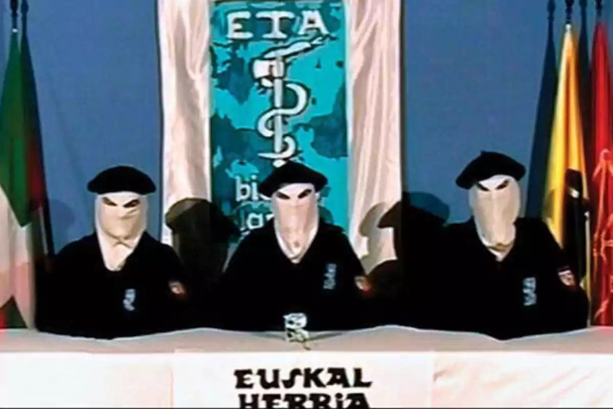 Tres personas enmascaradas con boinas negras sentadas en una mesa con un cartel que dice "Euskal Herria" y un fondo con el logotipo de ETA.