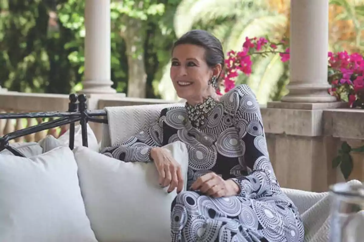 Una mujer sonriente con un vestido de patrones circulares en blanco y negro, sentada en un sofá al aire libre con cojines blancos, rodeada de columnas y flores rosas.