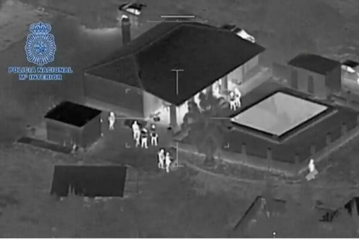 Imagen aérea en blanco y negro de una casa con varias personas alrededor, con el logotipo de la Policía Nacional y el Ministerio del Interior en la esquina superior izquierda.