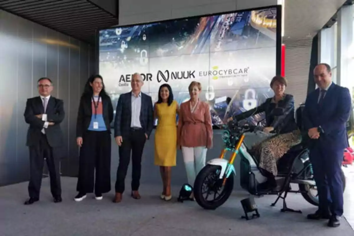 Un grupo de personas posando frente a una pantalla que muestra los logotipos de AENOR, NUUK y EUROCYBCAR, con una motocicleta eléctrica en primer plano.