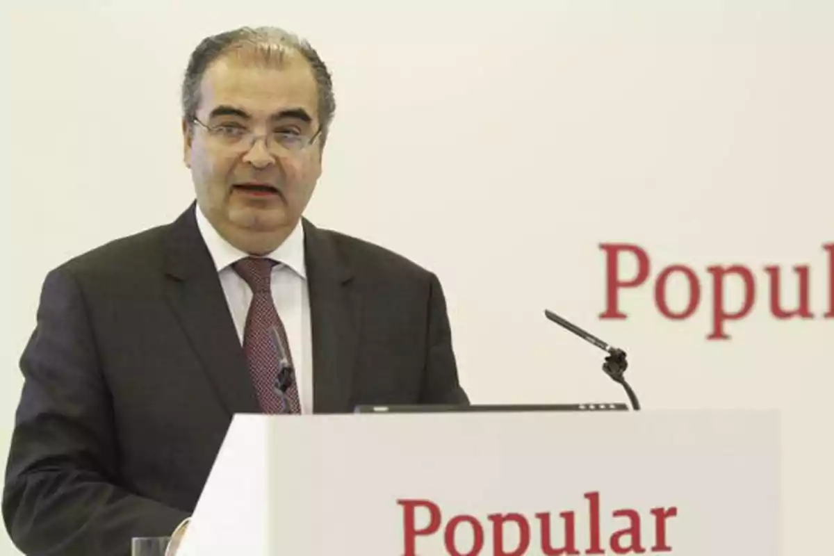 Hombre de traje y corbata hablando en un podio con el logo de "Popular" en el fondo.