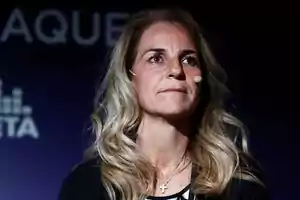 Una mujer con cabello rubio y ondulado, usando un micrófono de diadema y un collar con una cruz, en un escenario con fondo oscuro.