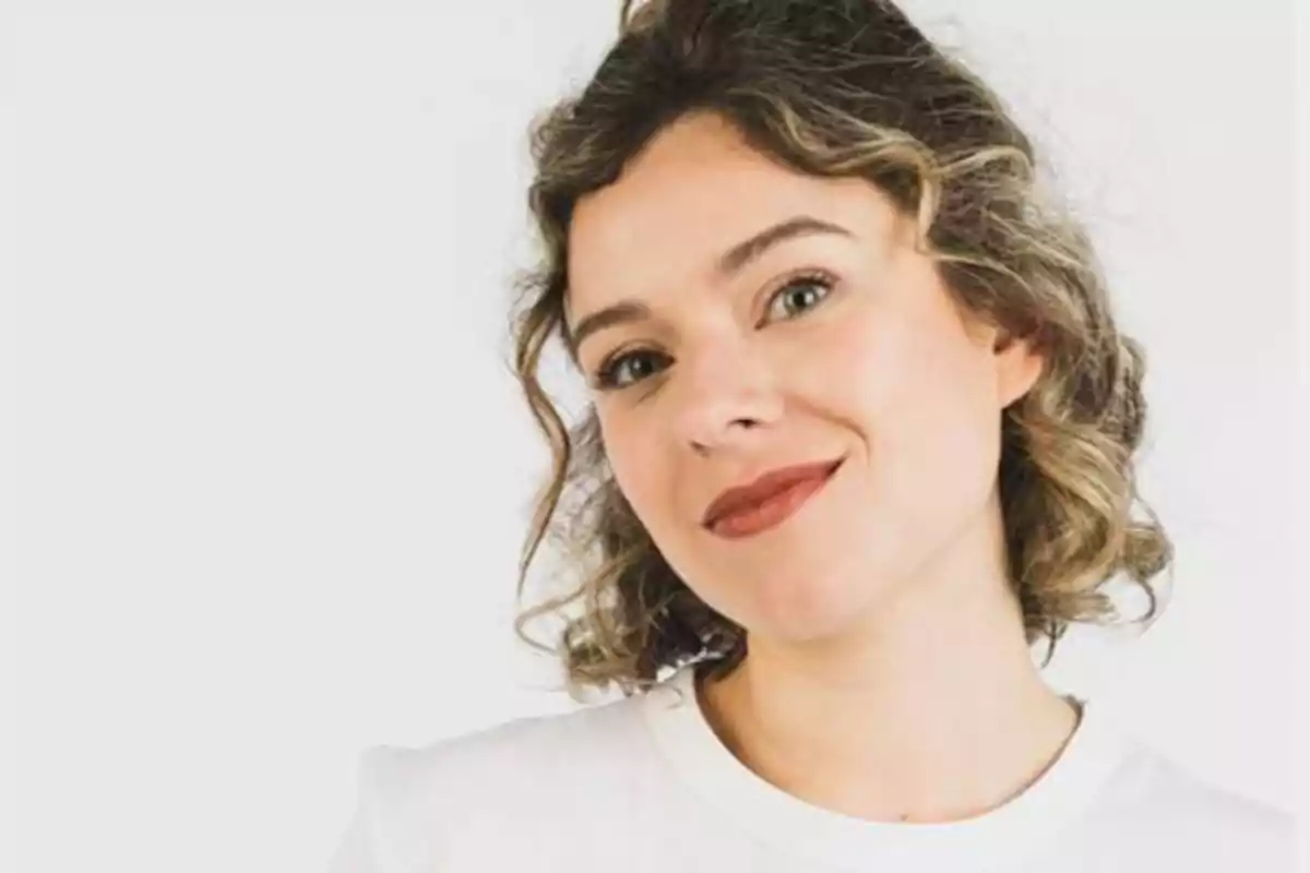 Mujer con cabello rizado y expresión sonriente, vestida con camiseta blanca sobre fondo blanco.