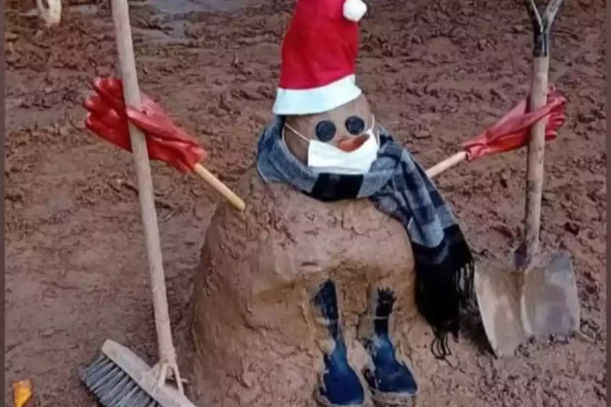 Un muñeco de barro con sombrero de Navidad, bufanda, mascarilla, guantes rojos y botas, sosteniendo una escoba y una pala.