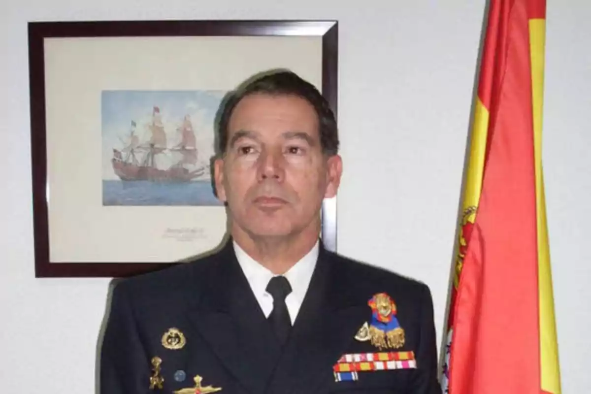 Un oficial militar en uniforme de gala posando junto a una bandera de España y un cuadro de un barco antiguo.