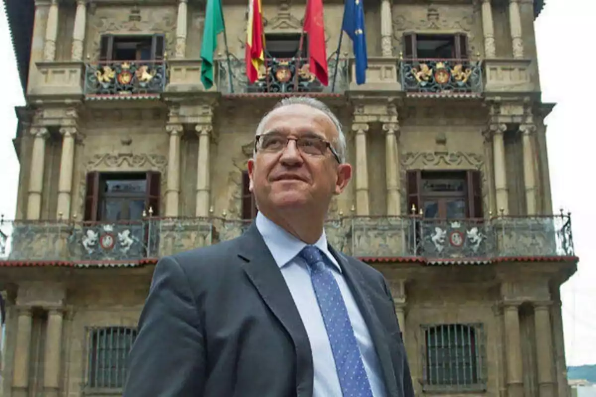 Hombre de traje y corbata frente a un edificio histórico con banderas.