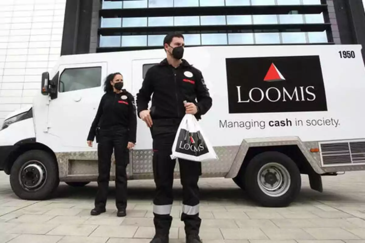 Dos personas con mascarillas y uniformes de seguridad están de pie junto a un vehículo blindado de la empresa Loomis.