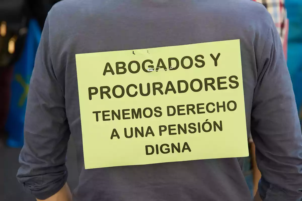 Persona vistiendo una camiseta gris con un cartel amarillo en la espalda que dice "Abogados y procuradores tenemos derecho a una pensión digna".