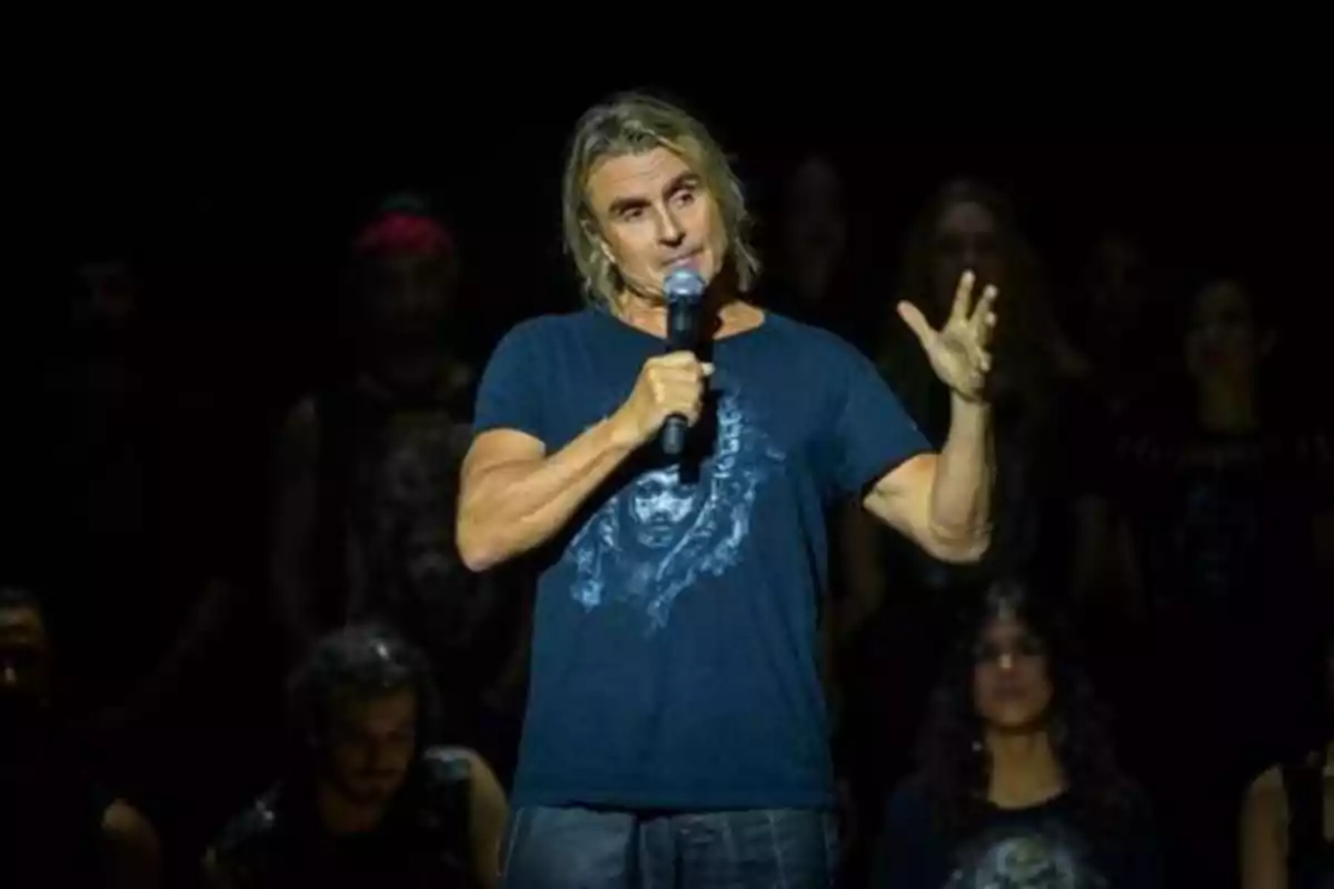 Hombre con camiseta azul hablando en un micrófono en un escenario oscuro.