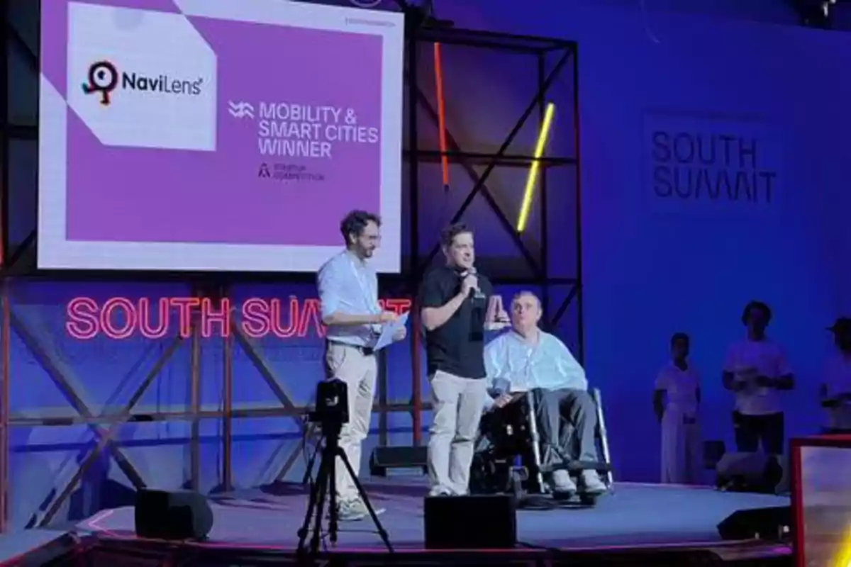 Tres personas están en un escenario durante un evento llamado "South Summit", una de ellas en silla de ruedas, mientras en la pantalla se muestra el logo de NaviLens y el texto "Mobility & Smart Cities Winner".