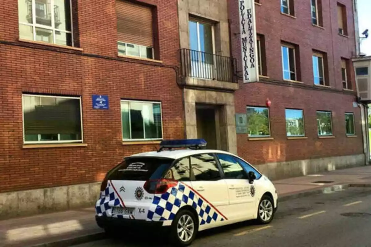 Un coche de la policía municipal está estacionado frente a un edificio de ladrillo rojo con varias ventanas y un cartel que dice "Policía Municipal Udaltzaingoa".
