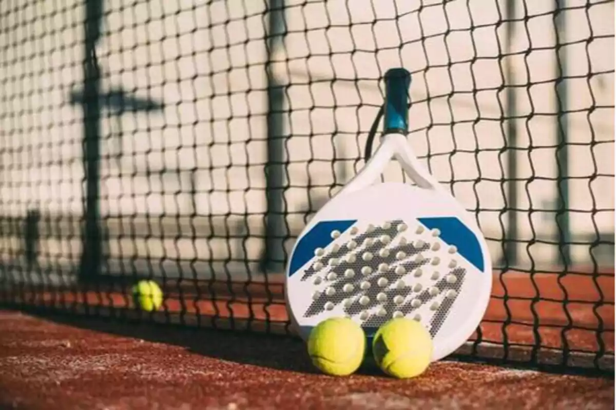 Raqueta de pádel blanca con detalles azules apoyada en una red, acompañada de dos pelotas amarillas sobre una cancha de arcilla.