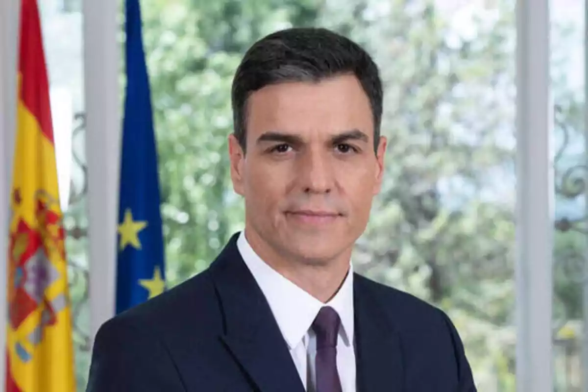 Hombre con traje oscuro y corbata frente a banderas de España y la Unión Europea.