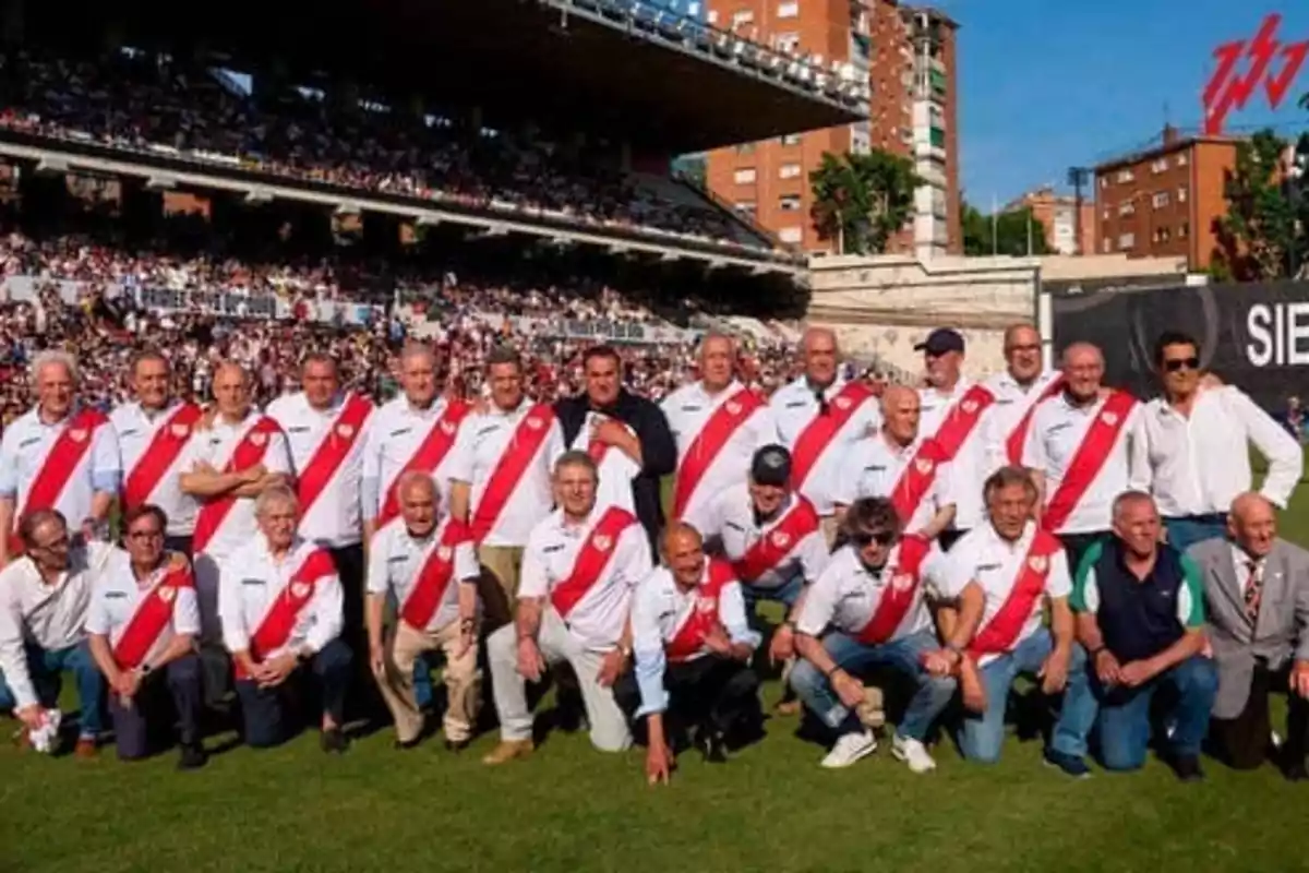 Un grupo de personas posando en un estadio de fútbol, todas vestidas con camisetas blancas con una franja roja diagonal.