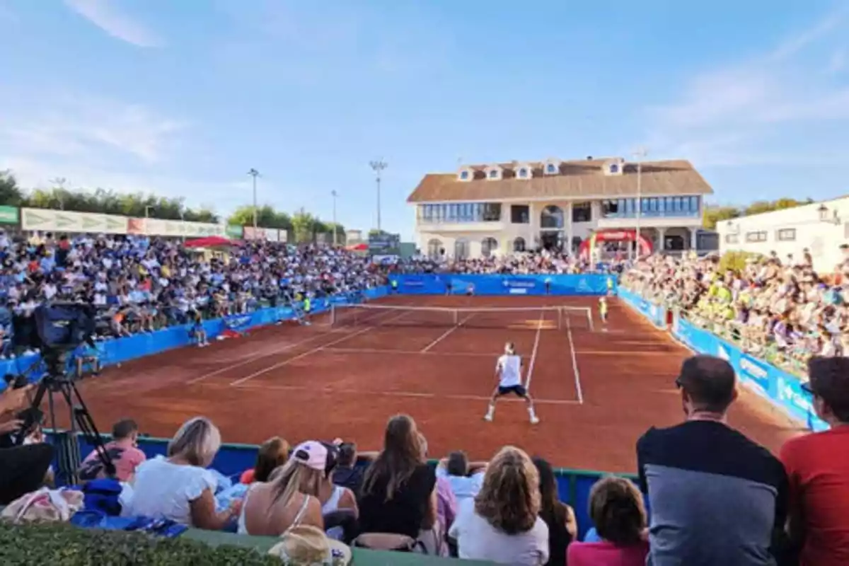 Partido de tenis en una cancha de tierra batida con una multitud de espectadores en las gradas y un edificio grande al fondo bajo un cielo despejado.