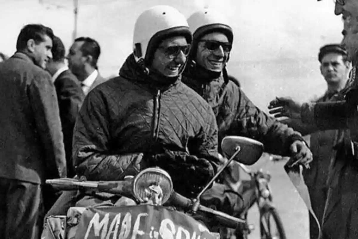 Dos motociclistas con cascos y gafas de sol sonríen mientras están en una motocicleta con un cartel que dice "MADE in SPAIN", rodeados de varias personas.